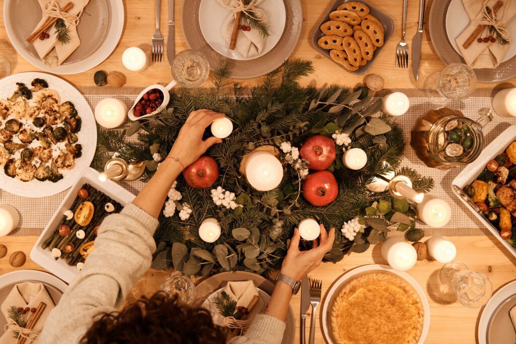 A table set for a festive holiday dinner complete with food, dishes, and glasses, with a person reaching in to place candles candles; highlighting holiday hacks, such as eating together, that can help relieve stress.